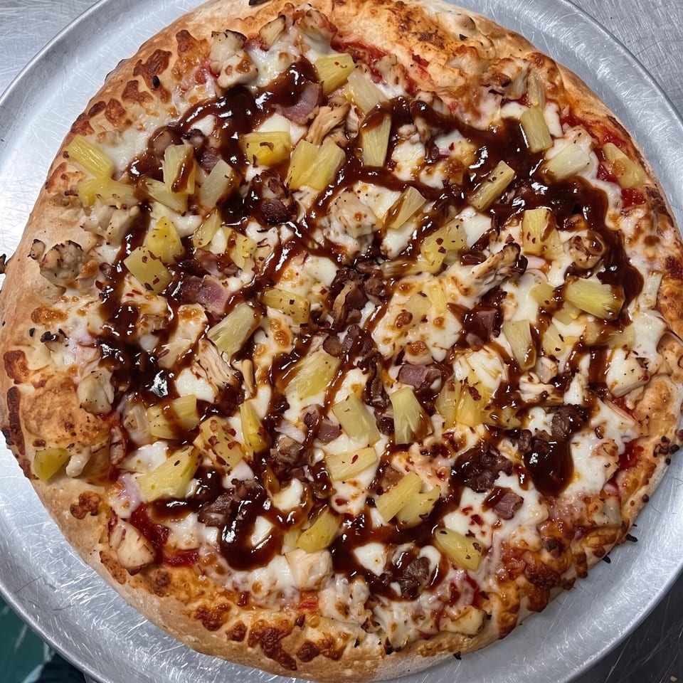 Golden-crusted pizza topped with melted cheese, chicken, pineapple, bacon, mushrooms, herbs, and glossy barbecue sauce.