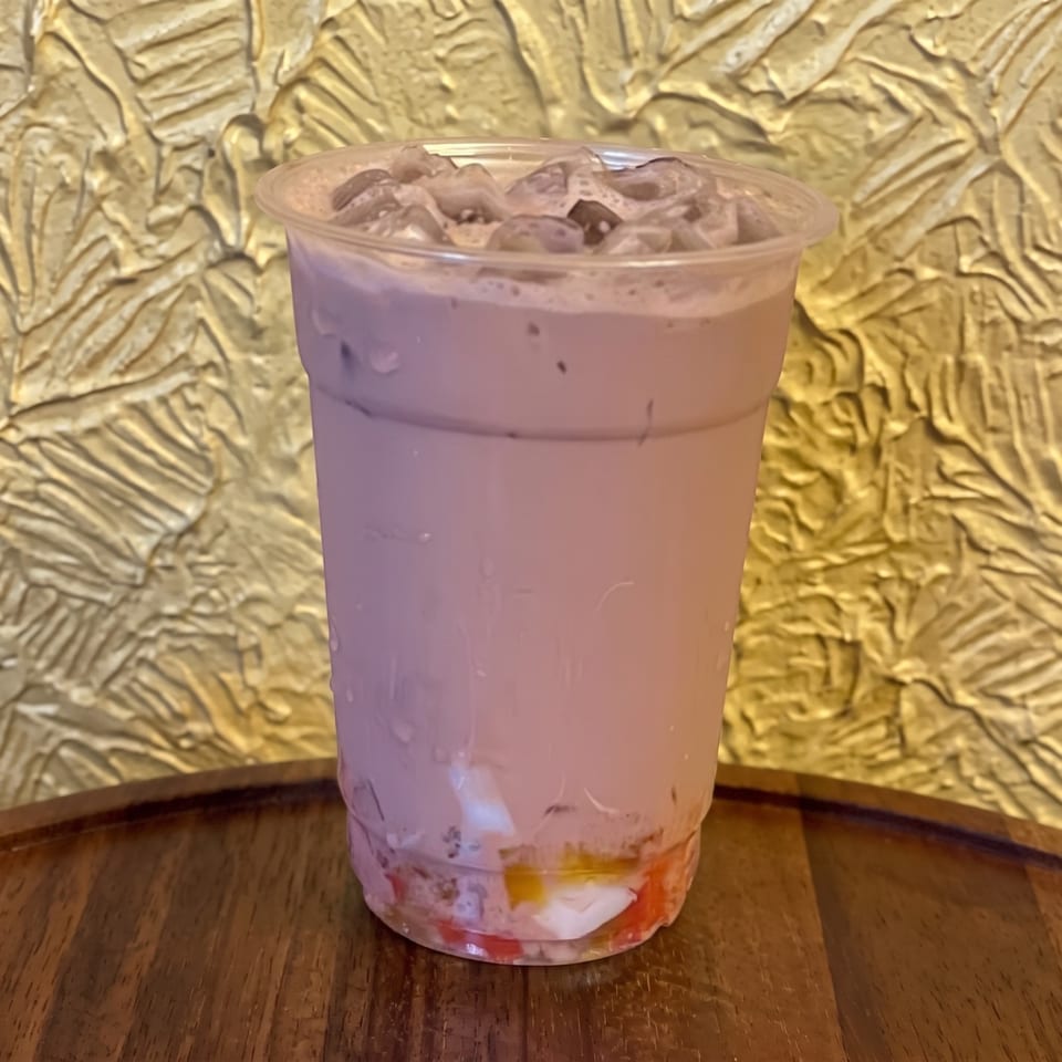 Creamy pink iced drink topped with fruit pieces in a clear cup on a wooden table against a gold botanical backdrop.